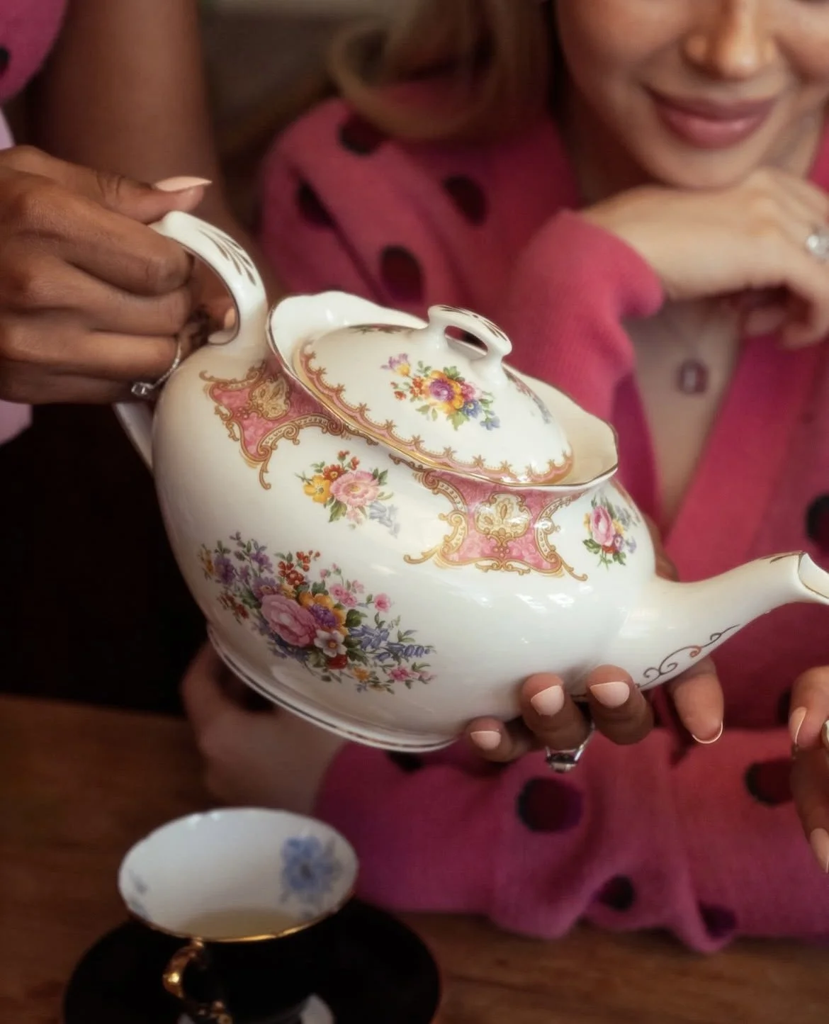 Close-up of two women holding a vintage floral teapot, with one pouring tea and the other smiling, wearing pink with black polka dots. A tea cup with floral design is on the table.