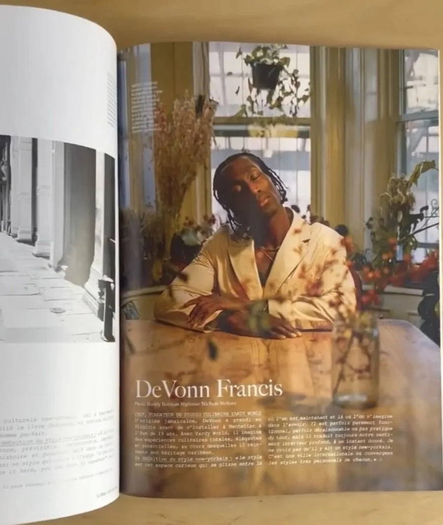A woman with short hair sitting at a table in a room with large windows, plants, and natural light.