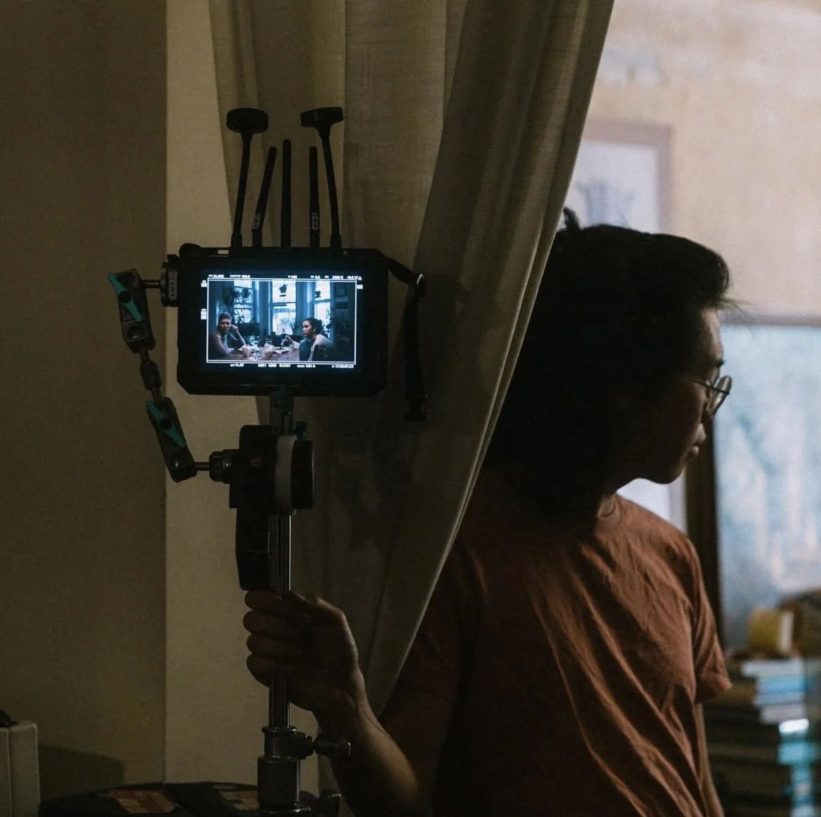 A woman with glasses holding a camera on a tripod, capturing a scene of two people sitting at a table in a room with large windows.