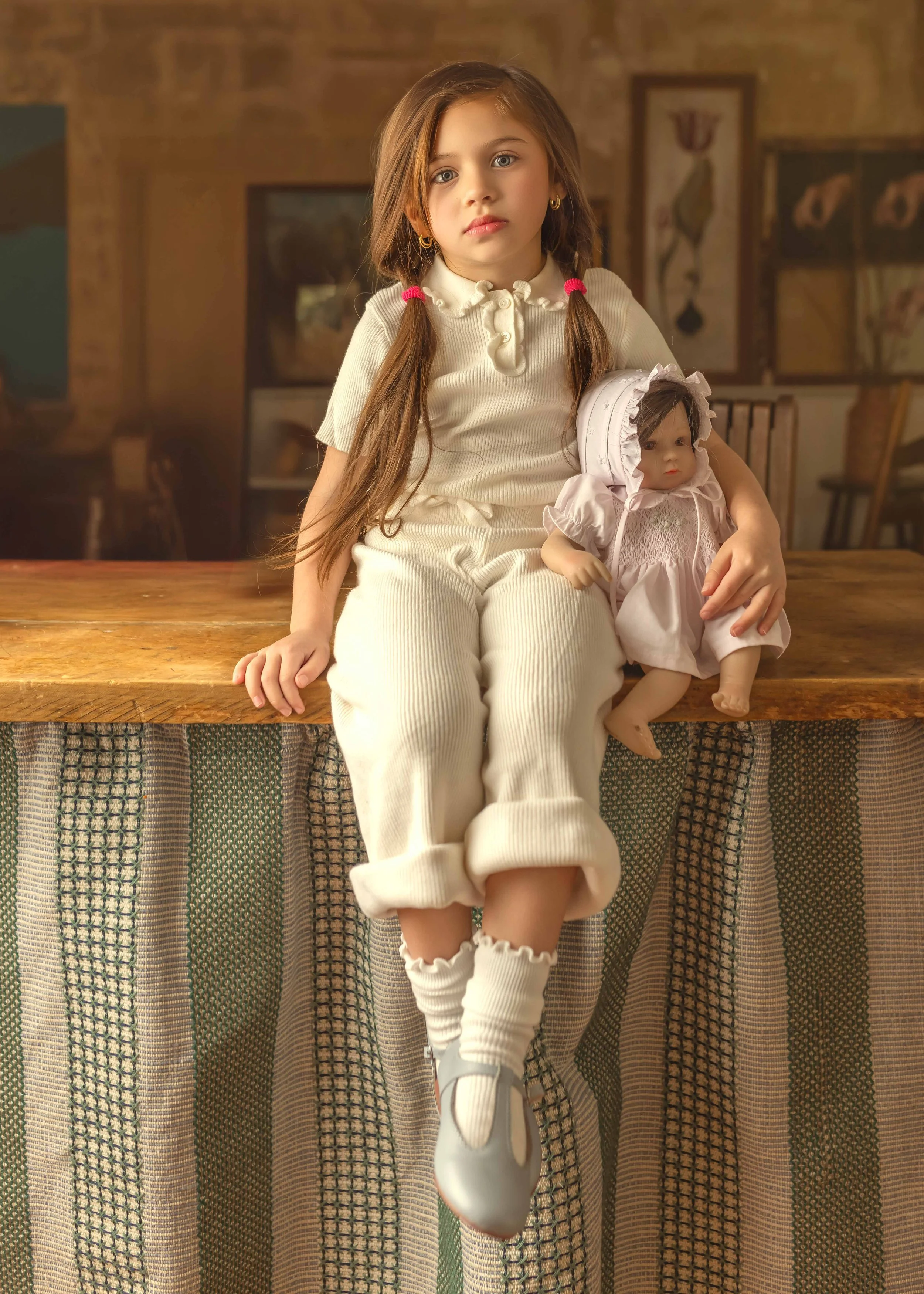 A young girl with long brown hair and blue eyes, wearing a cream-colored outfit, sitting on a wooden table holding a doll dressed in pink. The background shows a room with framed artwork on the walls.
