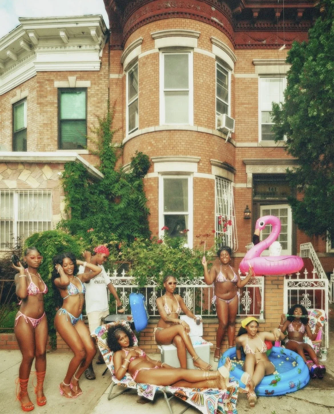 Group of women in bikinis enjoying a pool party outside a brownstone building with red brick and greenery.