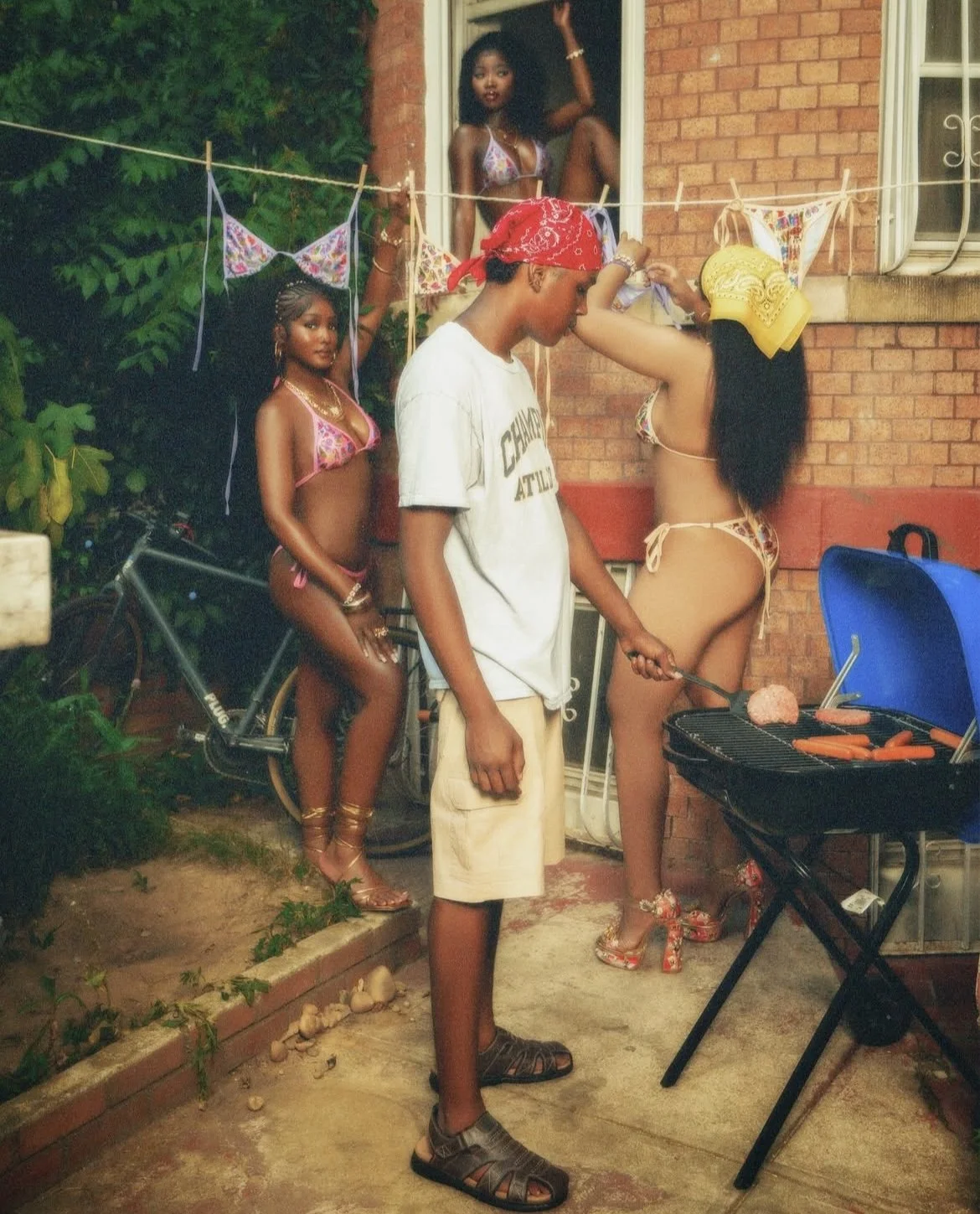 A group of four young women and a young man gathers outside a brick house. Three women are in bikinis, and one is in a yellow headscarf. One woman is cooking hot dogs on a small grill. The young man, wearing a white T-shirt, beige shorts, sandals, an
