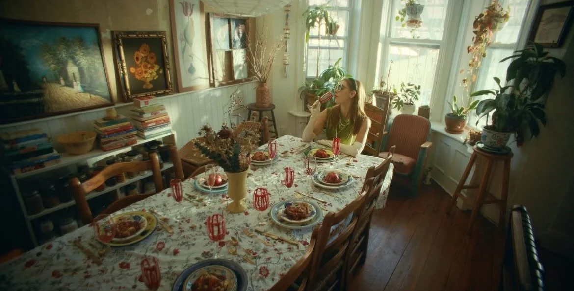 A woman sitting at a dining table sipping from a glass, surrounded by plates of food, with a cozy, well-lit room filled with flowers, paintings, and potted plants.