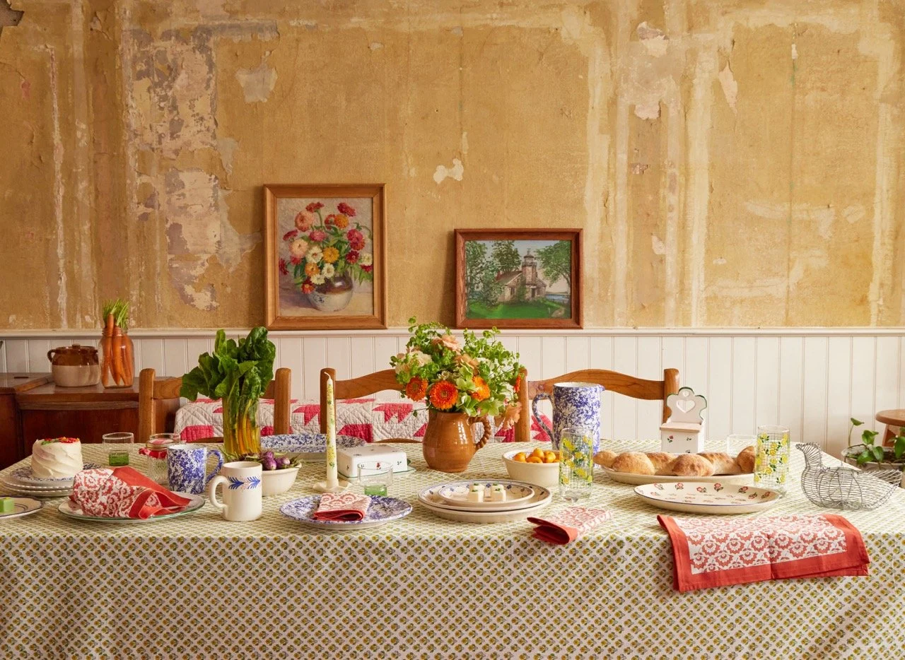 Cozy dining room with a long table set for a meal, decorated with flowers, plates, mugs, candles, and food, against a rustic yellow wall with two framed paintings.