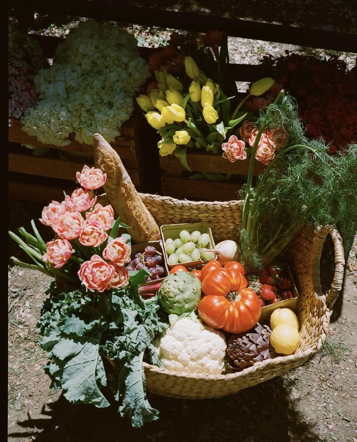 Basket of fresh vegetables and flowers, including cauliflower, tomatoes, zucchini, artichoke, strawberries, grapes, and pink tulips, with other flowers in the background.
