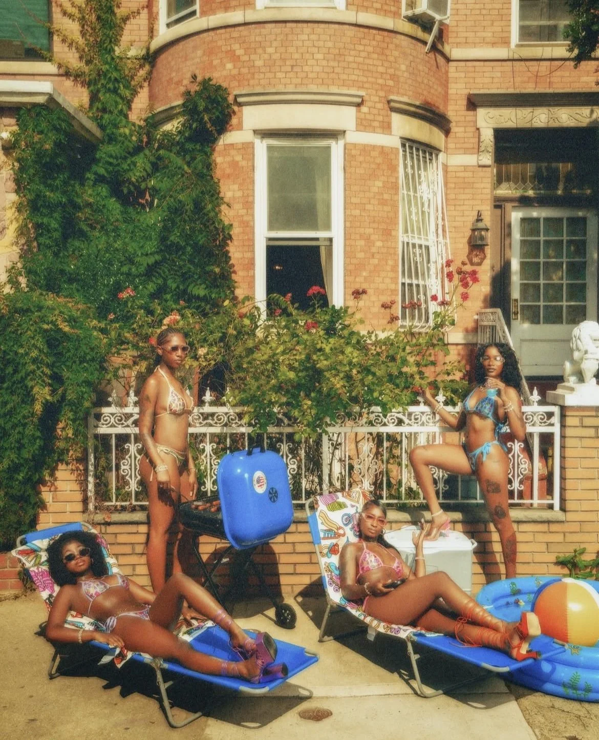 Four women relax outdoors in swimsuits on lounge chairs and a float, near a brick building with green plants and flowers, during daytime.