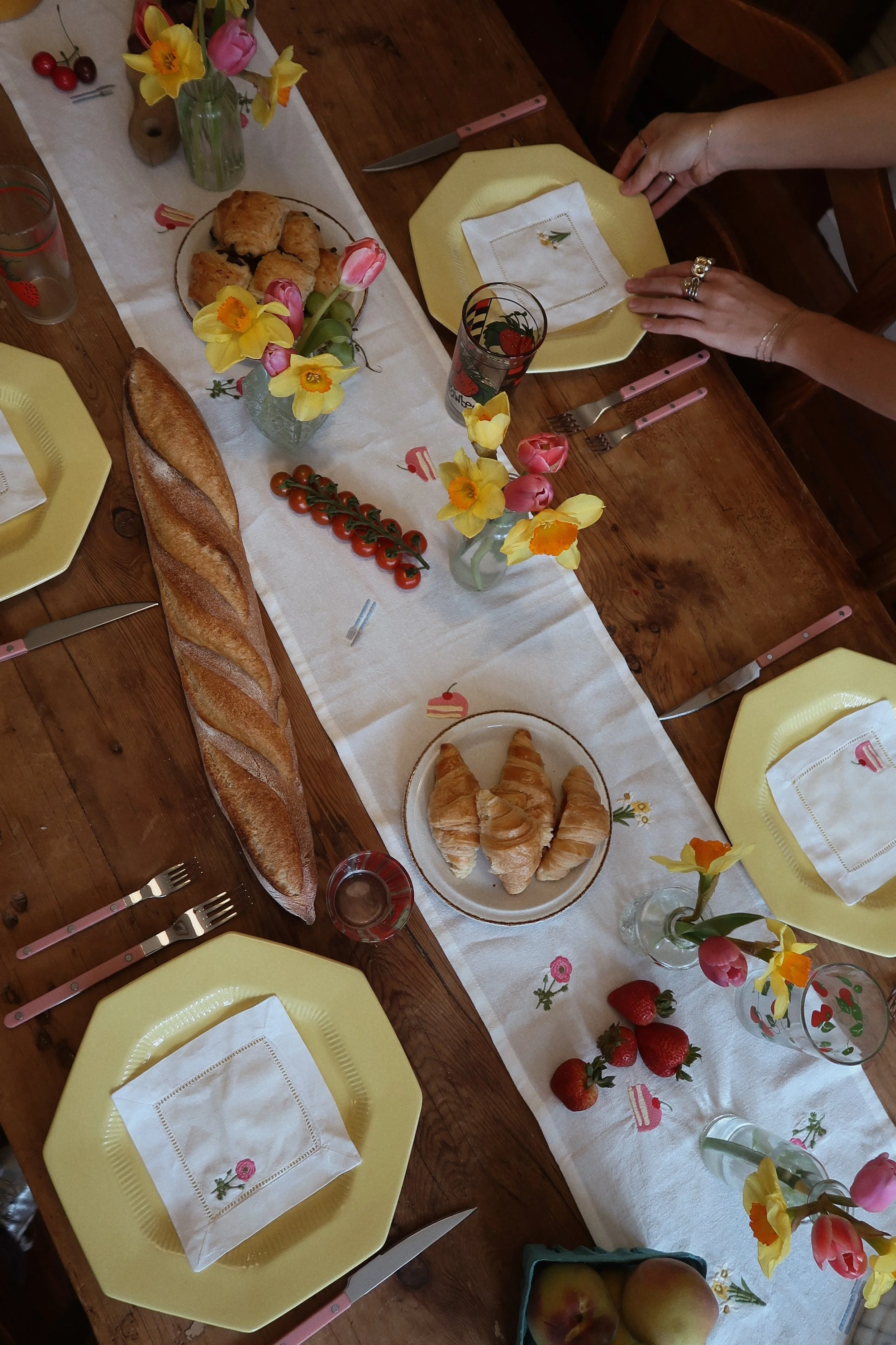 A breakfast table set with yellow plates, pink utensils, a long baguette, croissants, strawberries, cherry tomatoes, and flower arrangements with daffodils and tulips, all on a white table runner with cake and pastry illustrations.
