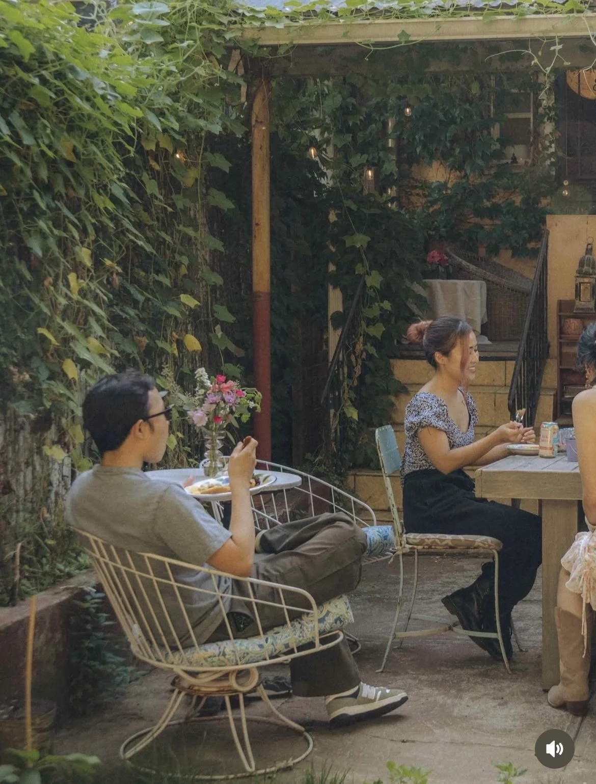 People sitting at a table in an outdoor garden dining area, surrounded by green vines and string lights, enjoying a meal and drinks.