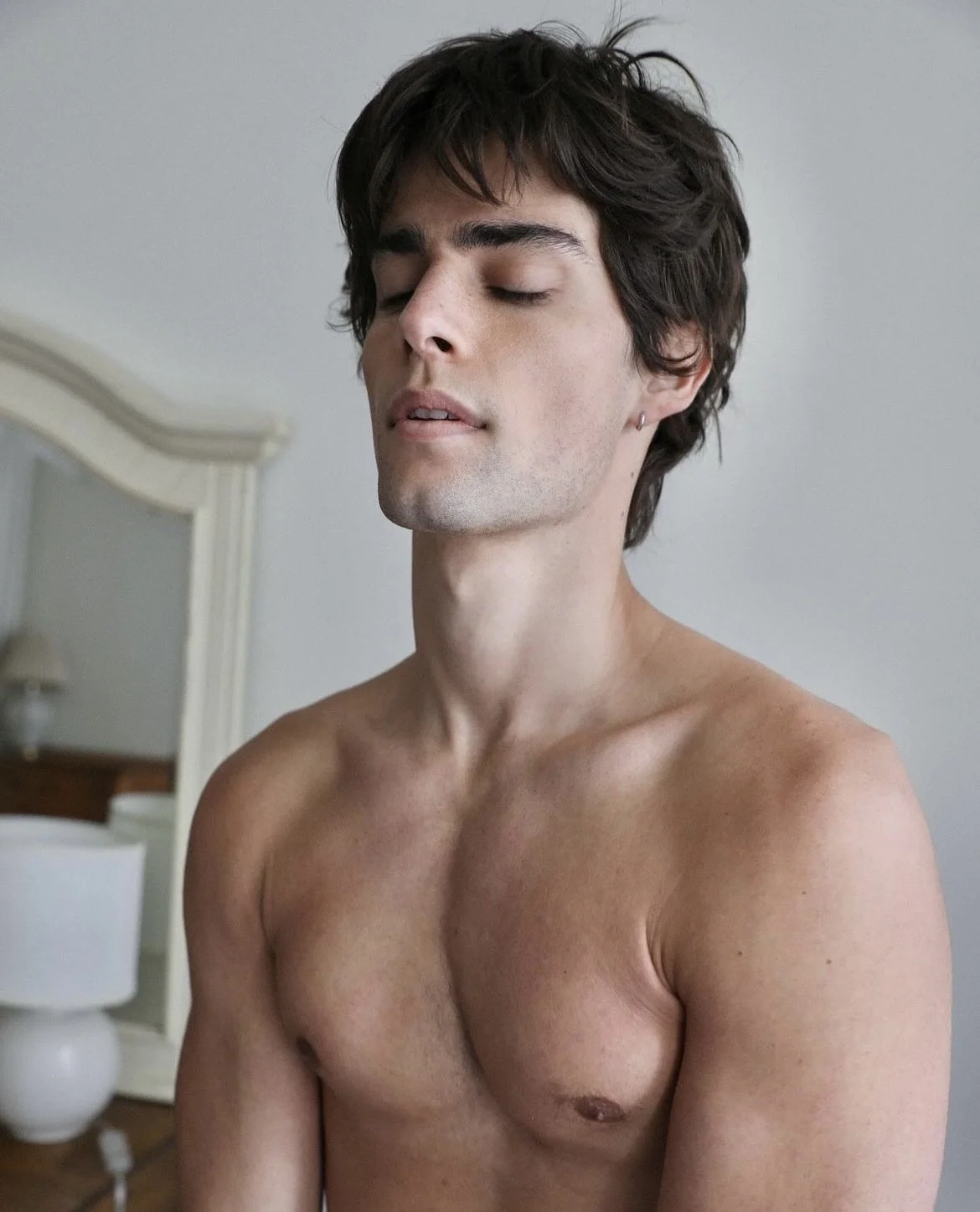 A shirtless young man with dark hair and closed eyes in a bedroom, with a white headboard and a nightstand with a lamp in the background.