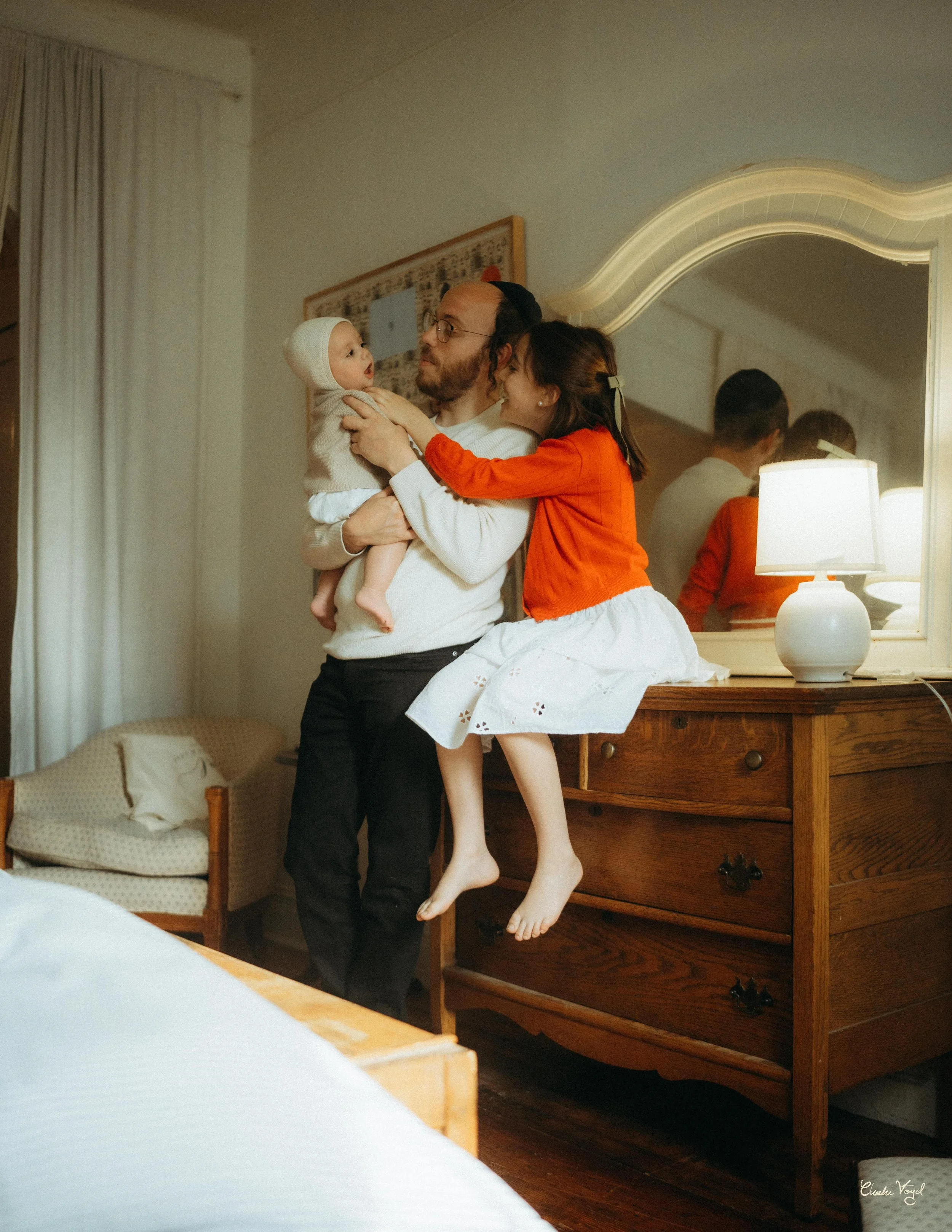 A family of three enjoying a moment together in a warmly lit room. The man is holding a baby, while a young girl in a red sweater and white skirt is sitting on a wooden dresser, reaching out to the baby. There is a mirror and a bedside lamp on the dr