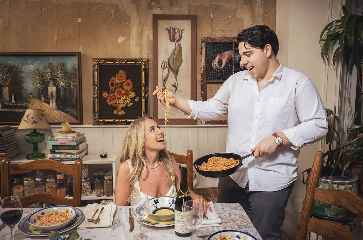 A man standing and holding a fork with spaghetti above a woman sitting at a dining table, smiling at him. The table is set with plates, glasses, and a bottle of wine. The background features paintings and a shelf with books and jars.