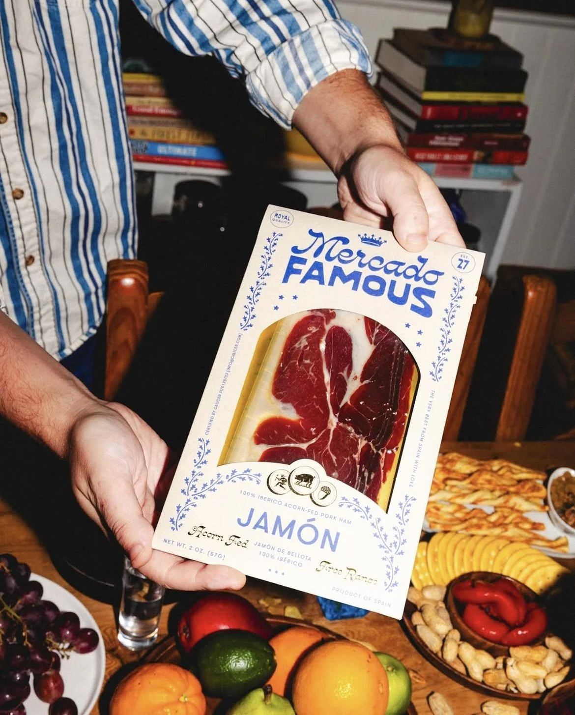 Person holding a package of Iberico acorn-fed pork ham called Jamón, labeled 'Mercado Famous', with a variety of fruits and snacks on the table in front of them.