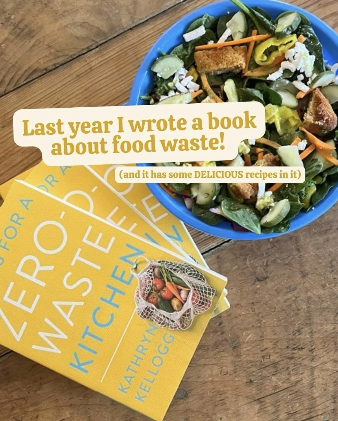 A blue bowl of salad with mixed greens, cherry tomatoes, shredded carrots, sliced cucumber, croutons, and shredded cheese on a wooden table. Next to it are two yellow books with the title "WASTE-FREE KITCHEN" and a miniature bag of vegetables on top.