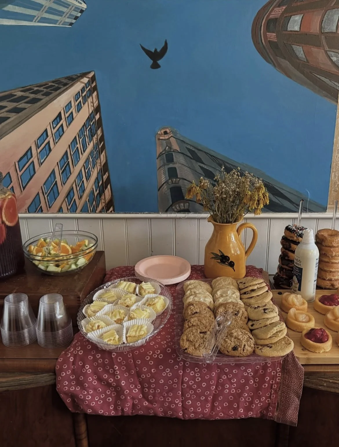 A table with baked goods including cookies, pastries, and scones, along with bowls of fruit salad, cups, a vase with dried flowers, and a mural of tall city buildings and a bird in flight overhead.