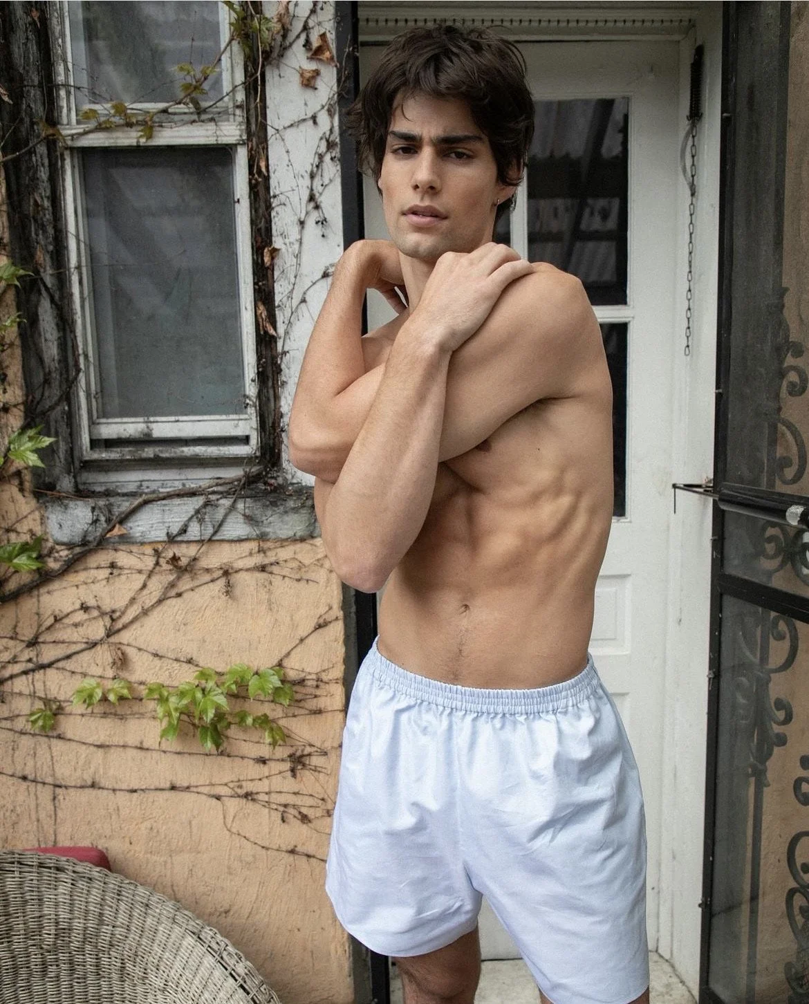 A young man with dark hair and a serious expression, shirtless, wearing white shorts, standing outside in front of a house with ivy and a window.
