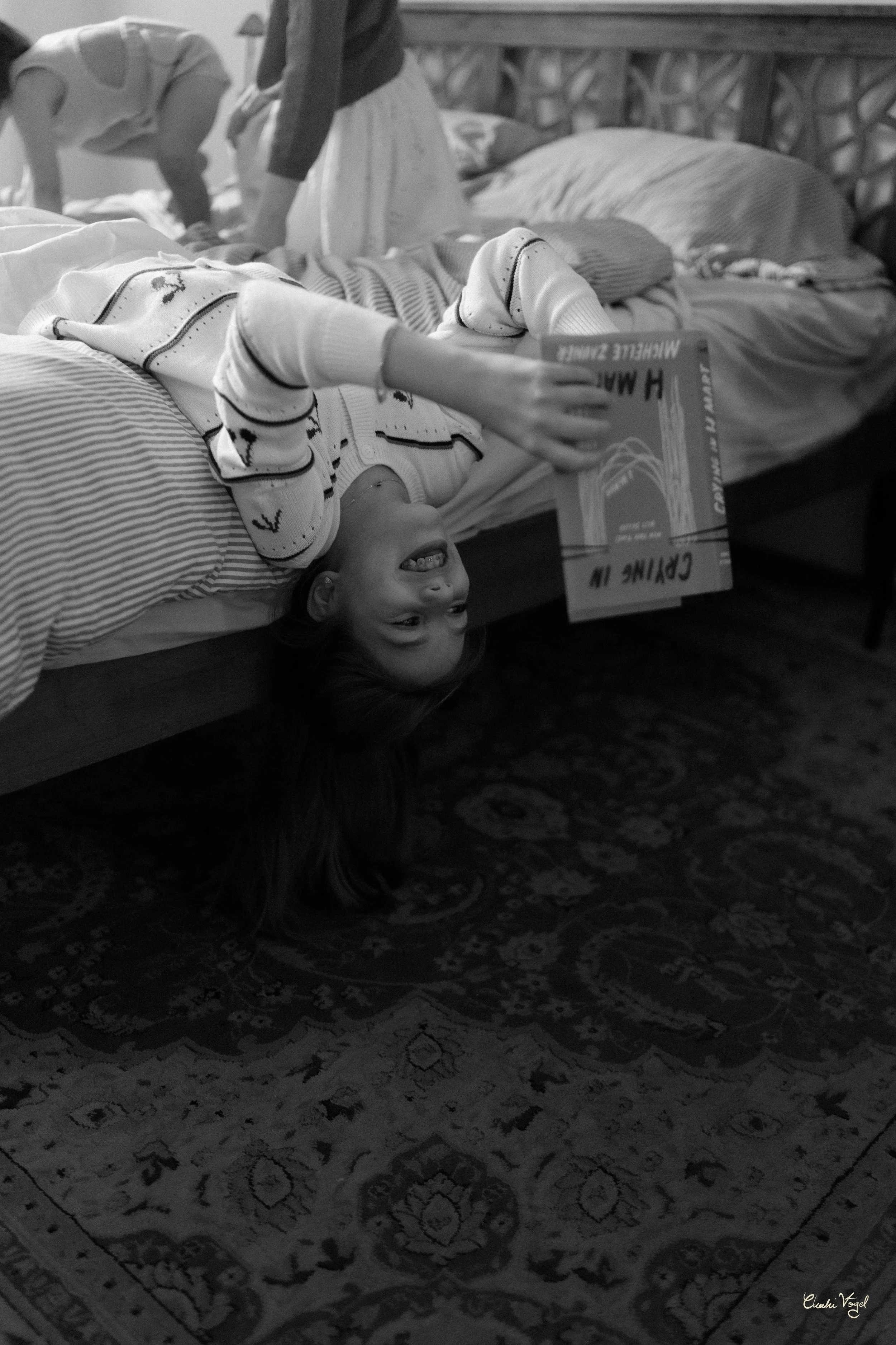 A young girl lies upside down on a bed, smiling while looking at a book she is holding. In the background, a child is visible, possibly playing or crawling.