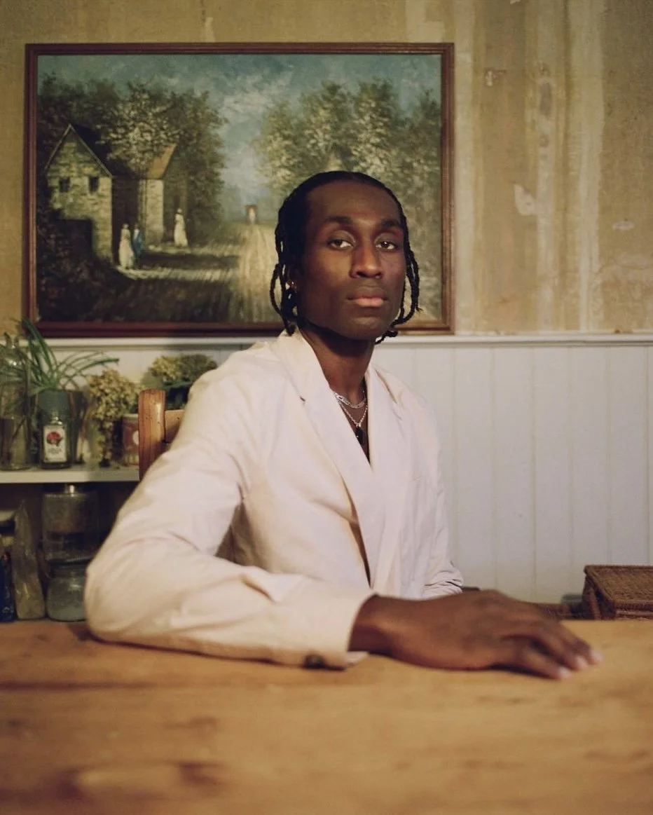 A man with braided hair and dark skin sitting at a wooden table, wearing a light-colored blazer, with a framed landscape painting featuring trees and a house on the wall behind him, and various plants and jars on a shelf to his left.