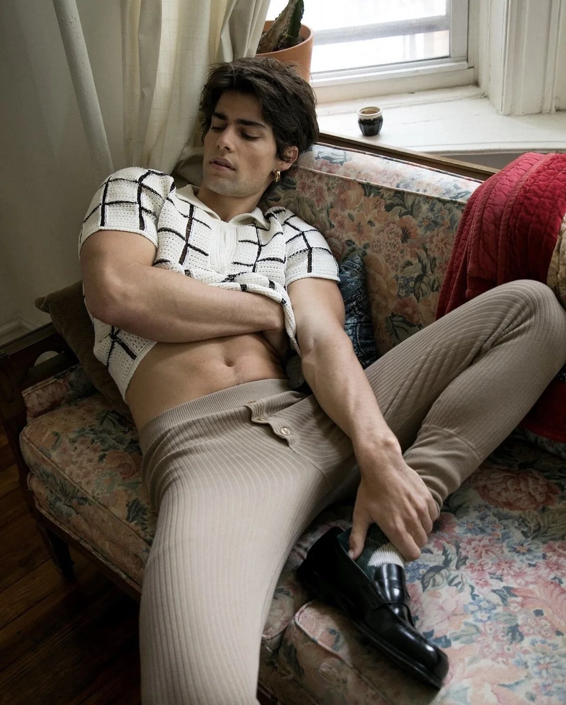 A young man with dark hair reclining on a floral sofa, wearing beige pants, a patterned shirt, and holding a black shoe in his hand. He appears to be sleeping or resting.