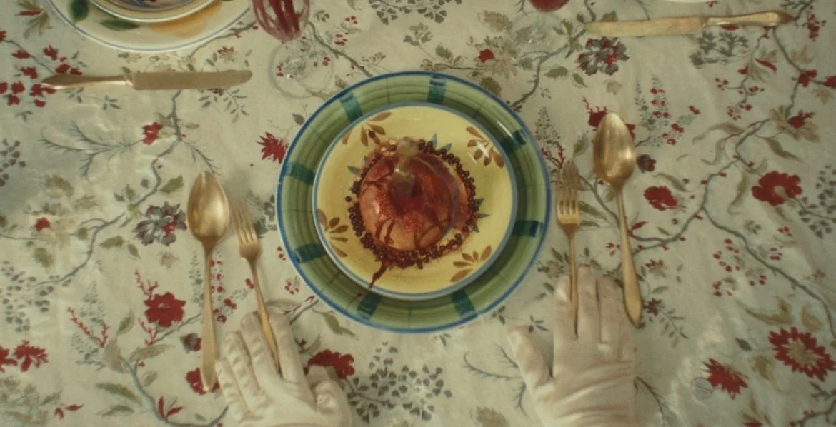 A table set for a meal with a floral tablecloth, disposable gloves, and a plate with a chocolate dessert topped with a cherry and drizzle, surrounded by matching bowls, forks, and spoons.