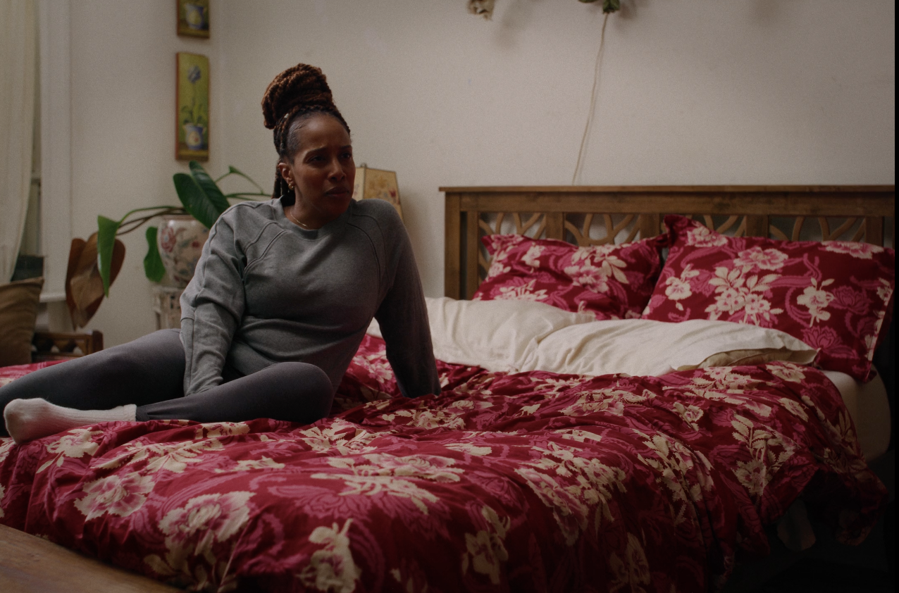 A woman with braided hair sitting on a bed with red floral bedding, wearing a gray sweatshirt and dark leggings, looking concerned or confused.