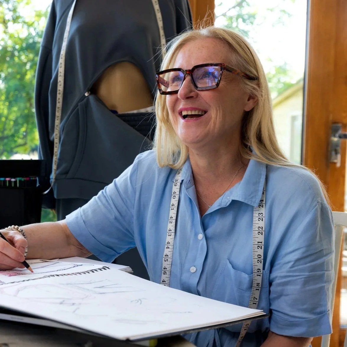 Joanne Nicholson with blonde hair, wearing glasses and a light blue shirt, smiling while drawing fashion designs for her adaptive medical clothing brand, EmAble.