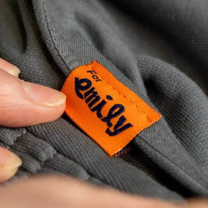 Close-up of an orange clothing label with dark blue text that reads "For Emily" sewn into a grey fabric garment.