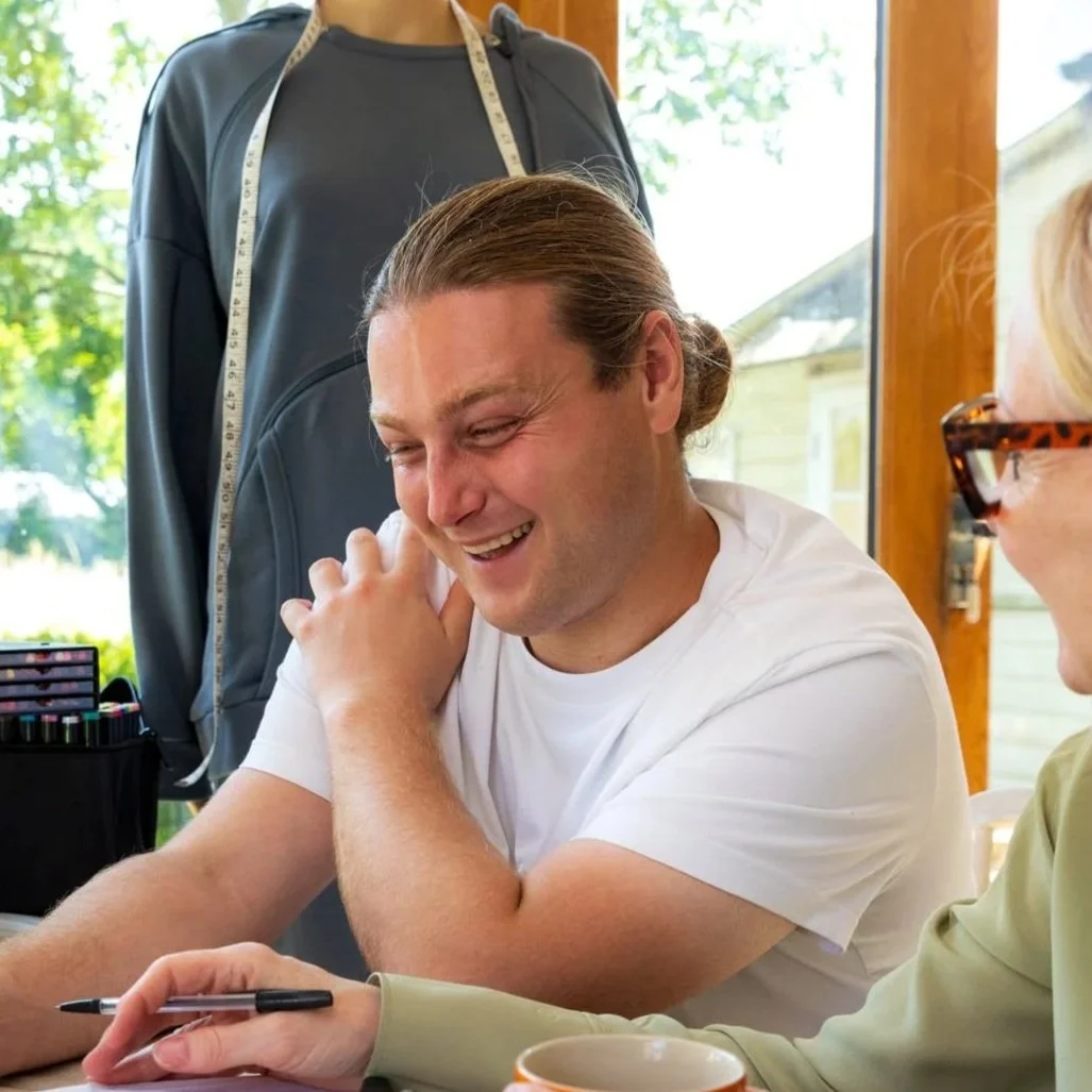 Matthew Rhodes, with long hair tied back, wearing a white t-shirt, is laughing while sitting at a table.