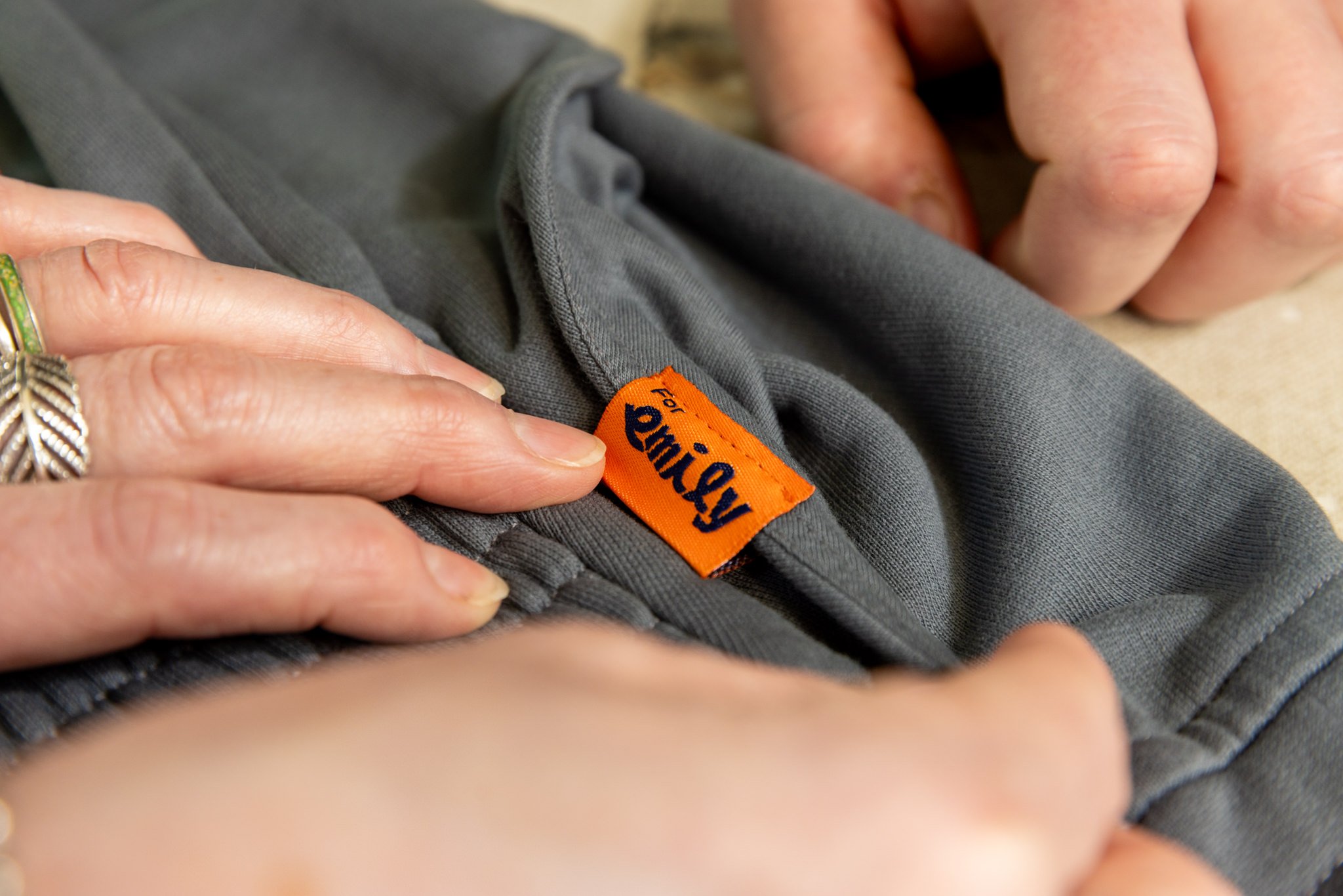Close-up of a person's hands pinching the waistband of grey clothing, with a bright orange tag labeled 'For Emily' visible.