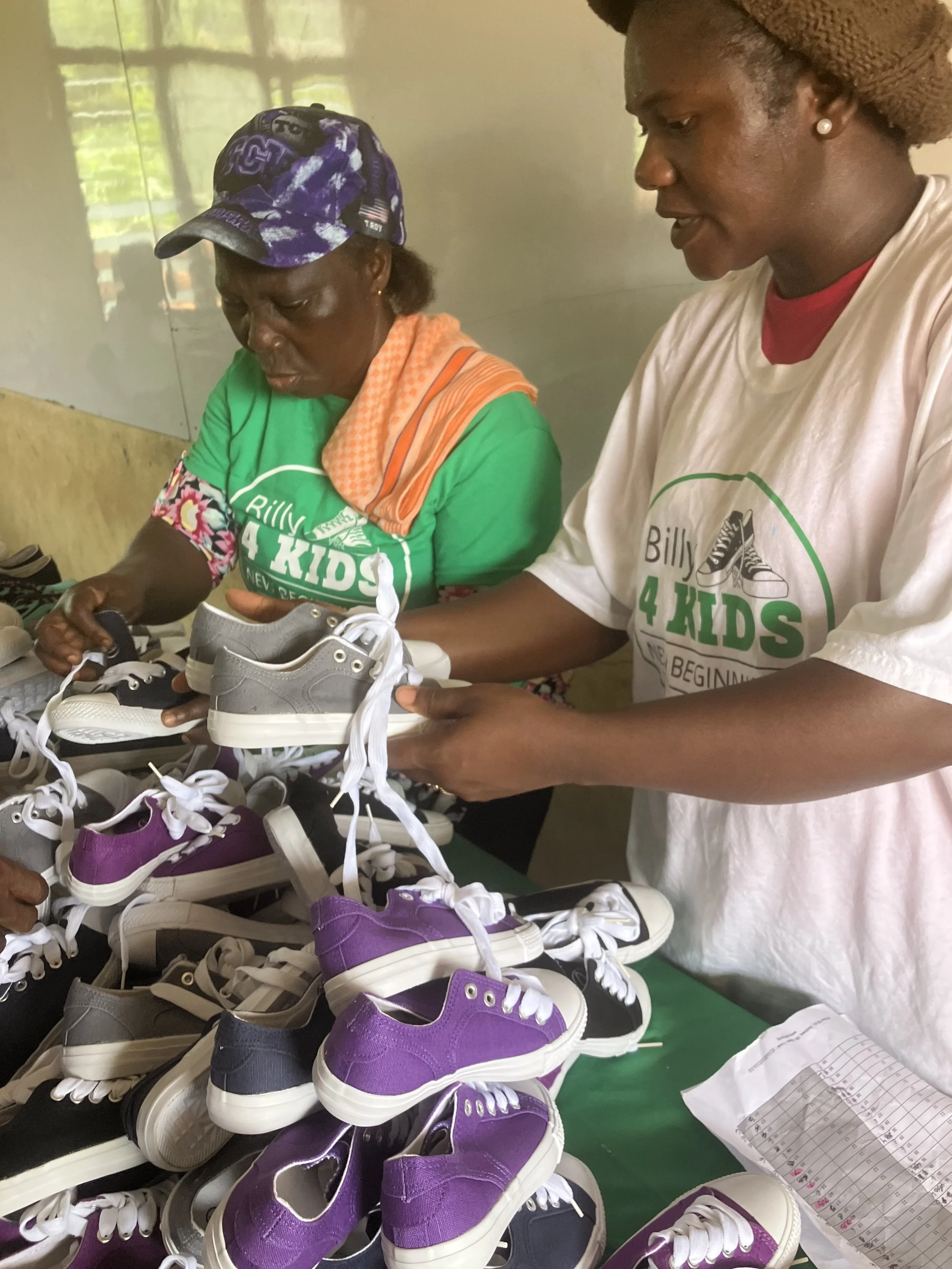 Two women sorting colorful children’s sneakers—including purple, black, grey, and white—on a table during a Billy4Kids donation event, in a room with a window reflecting outdoor trees.