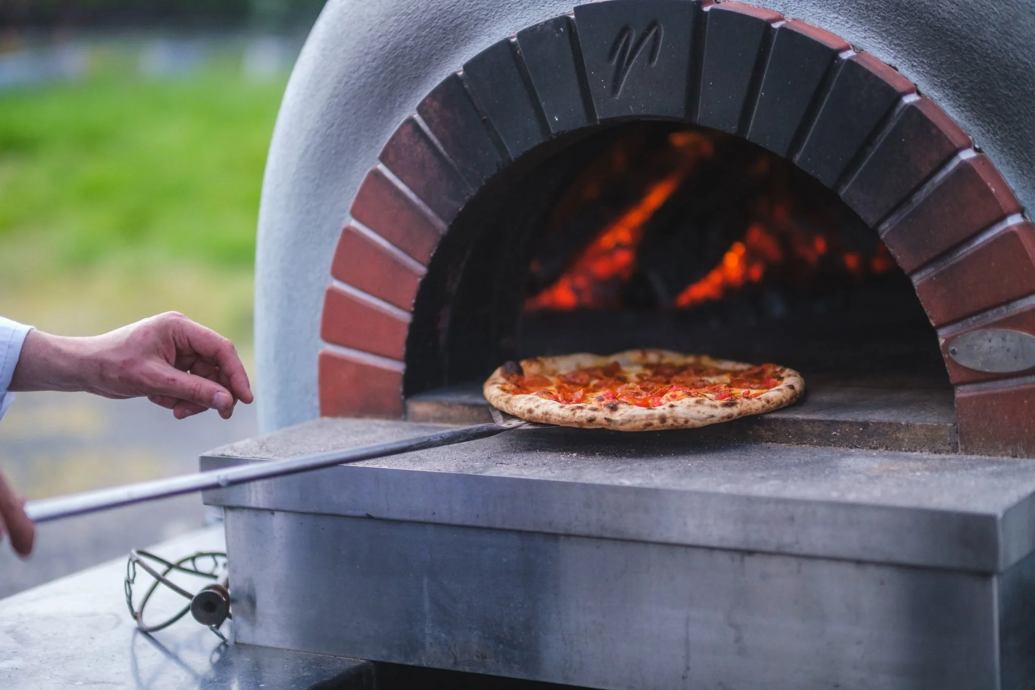A person placing a pizza into a wood-fired pizza oven with a burning fire inside.