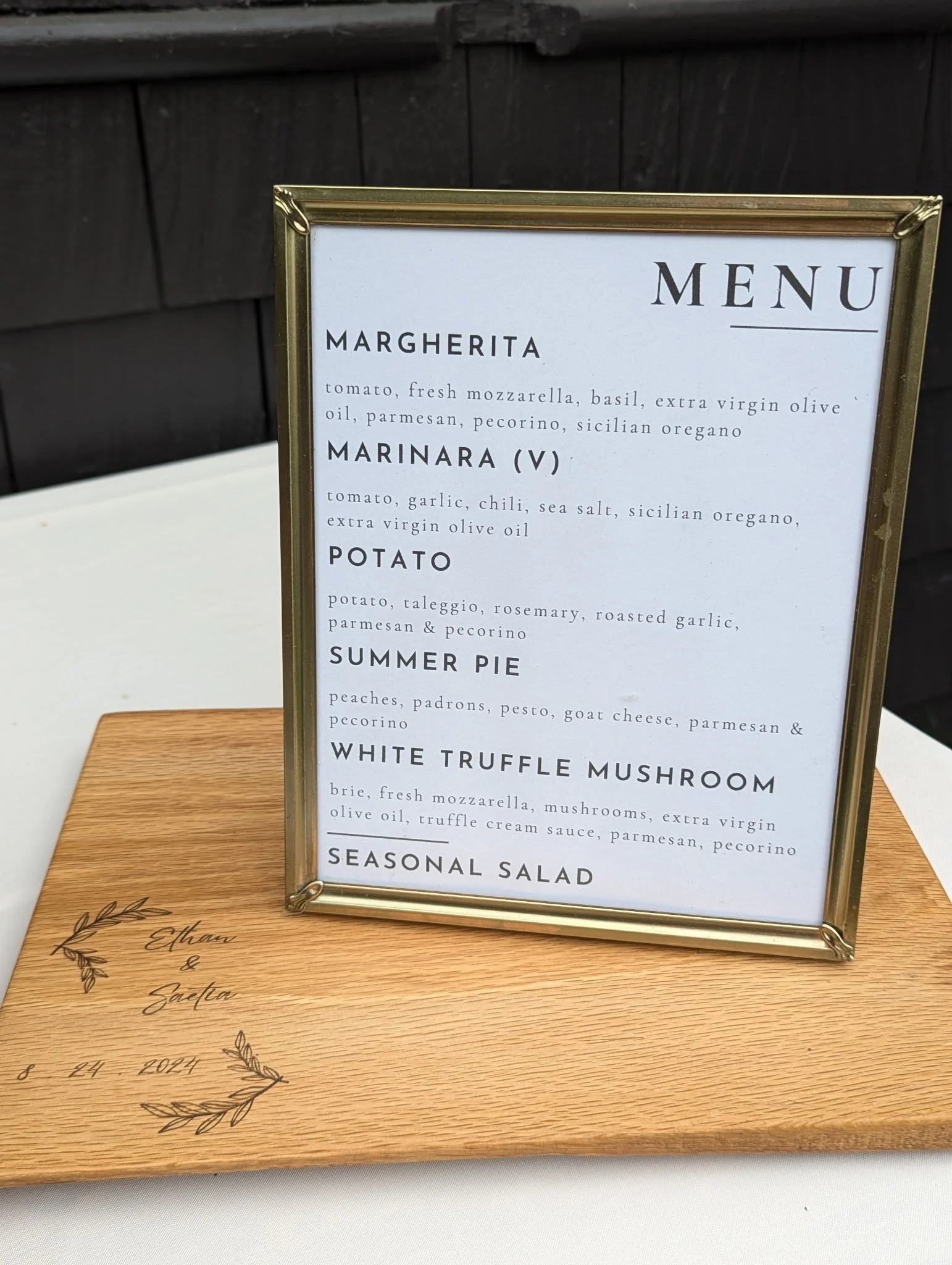 Restaurant menu featuring Margherita pizza, Marinara pizza, potato dish, summer pie, white truffle mushroom, and seasonal salad. The menu is displayed on a white paper in a gold frame, with a wooden base inscribed with the names Ethan and Saetra and a date.