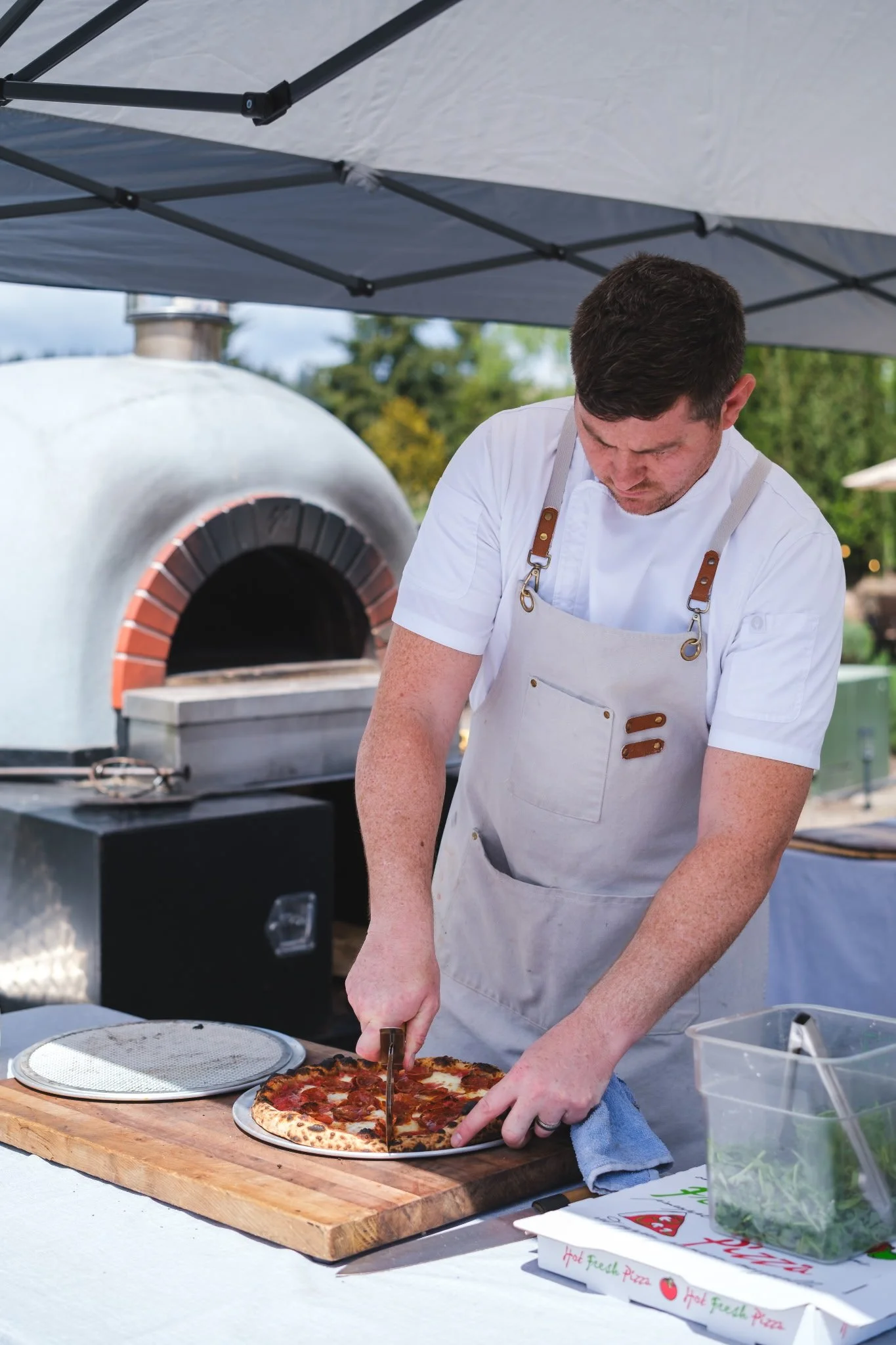 A man in a white shirt and beige apron slicing a pepperoni pizza on a wooden cutting board outdoors, with a wood-fired pizza oven in the background under an umbrella.