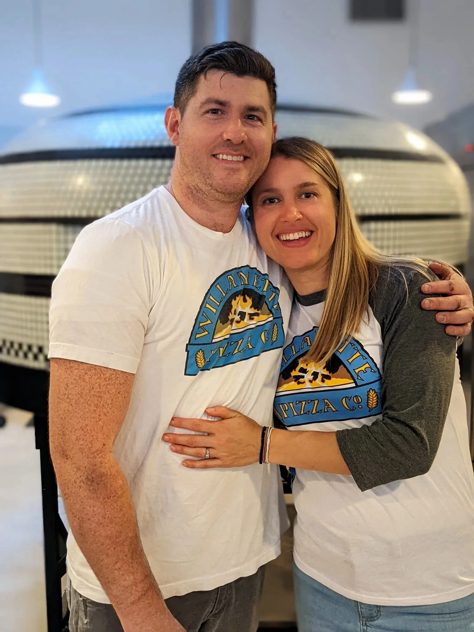 A smiling man and woman wearing matching Pizza Co. t-shirts, hugging each other inside a pizza restaurant with a pizza oven in the background.