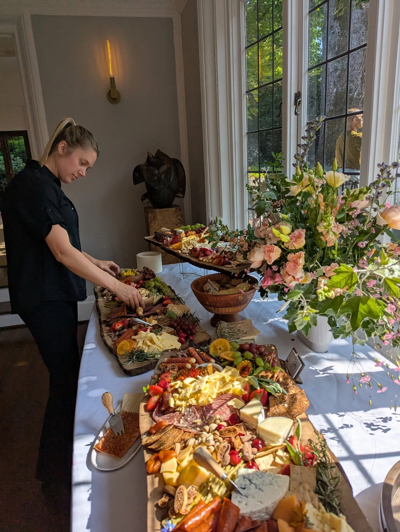 A woman arranging a large spread of cheese, meats, fruits, and other appetizers on a long table with a white tablecloth, near large windows with flower arrangements outside.