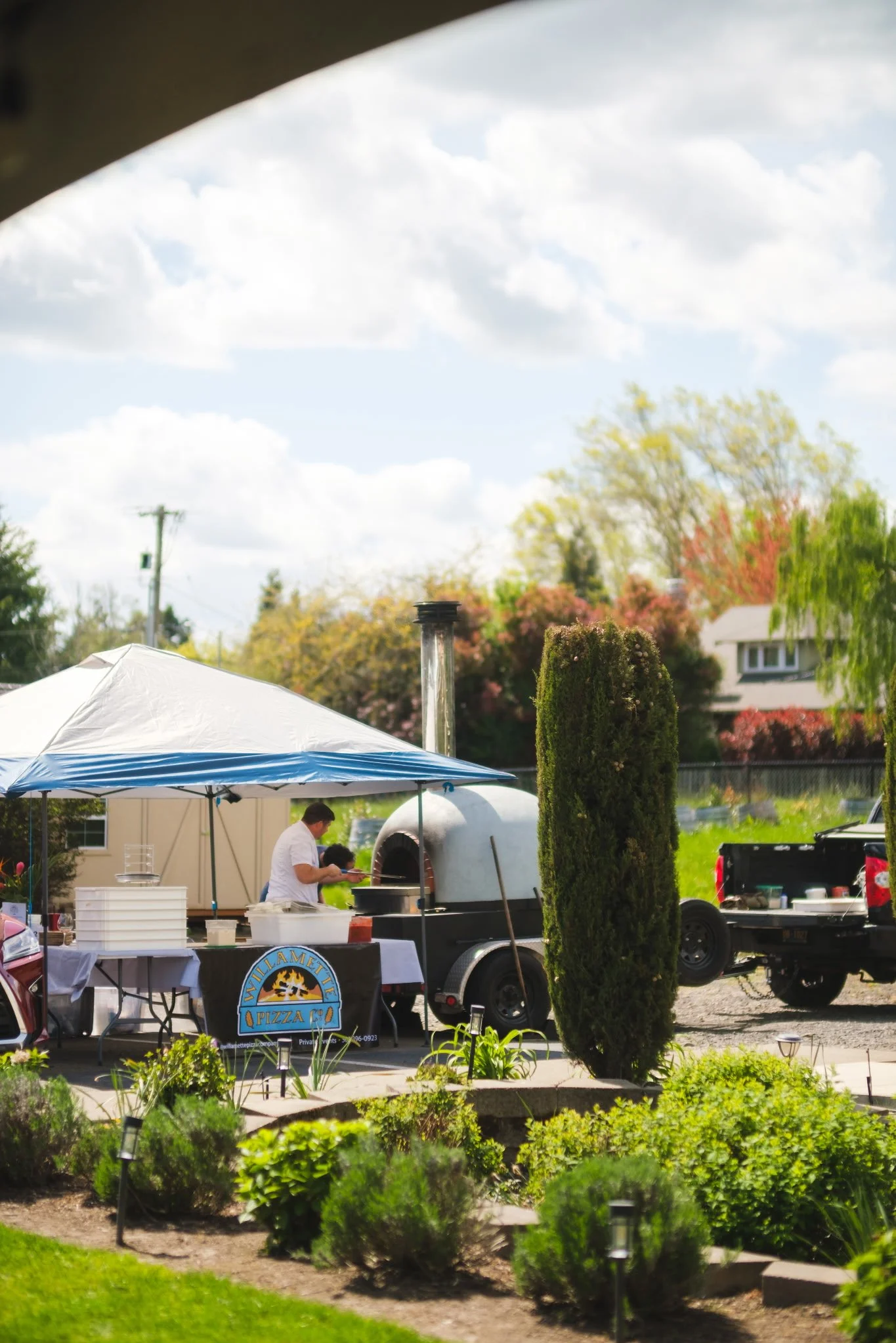 A man preparing pizza at a food truck with a wood-fired oven under a white and blue tent, in a lush green garden with shrubs and trees in the background.