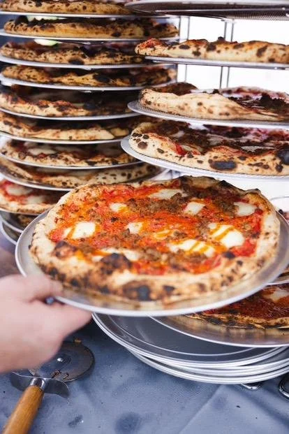 Multiple pizzas stacked on a cooling rack, with one pizza on a plate in the foreground being taken out.