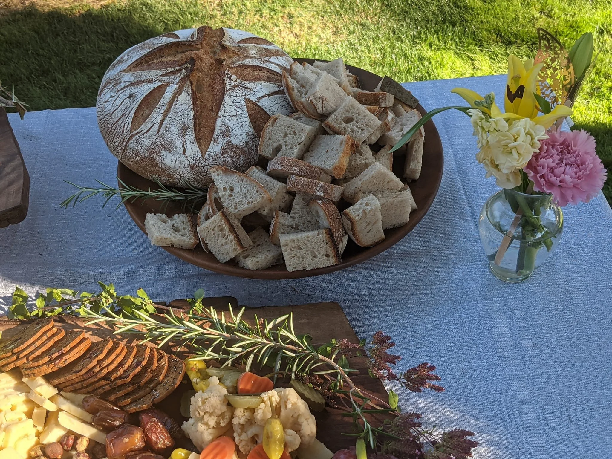 A picnic table with a loaf of bread and sliced bread on a wooden plate, a variety of sliced meats and cheeses, and a small glass vase with flowers including yellow lilies, pink carnations, and white and yellow flowers. The tablecloth is light blue, a
