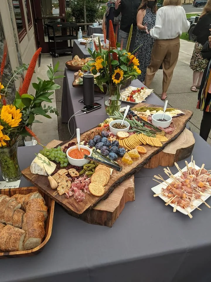 A charcuterie board with cheese, grapes, blueberries, dried fruit, crackers, and spreads on a wooden serving board. There are flower arrangements with sunflowers on the table, which has a gray tablecloth. People are gathered around in the background.