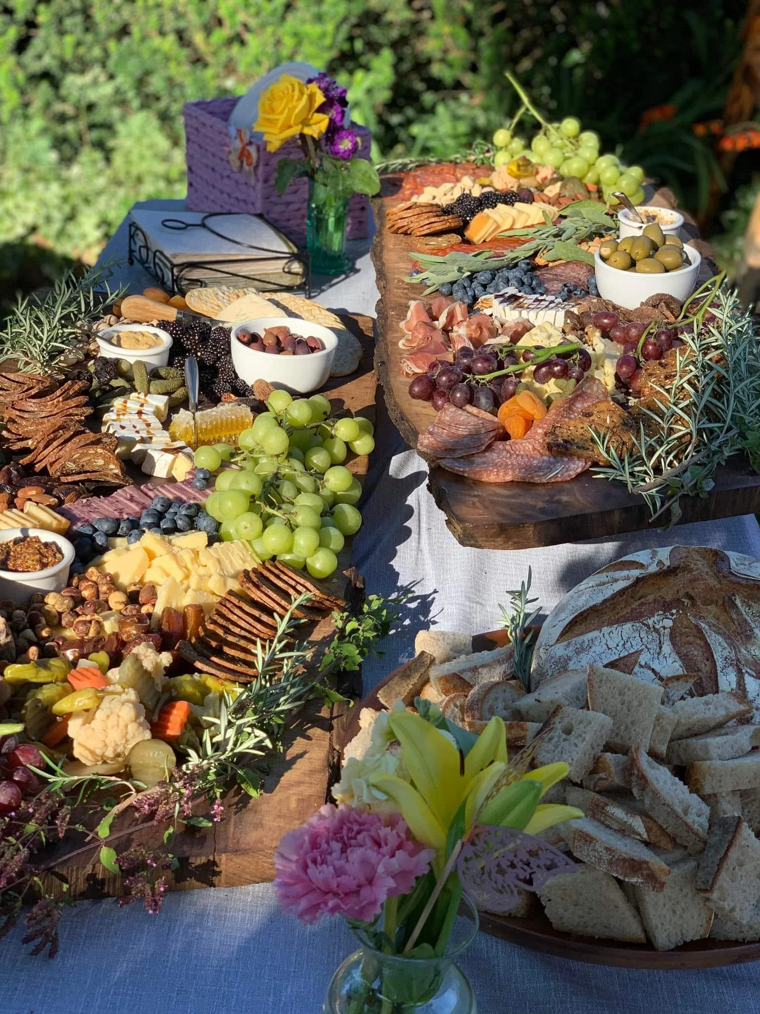 An outdoor charcuterie and cheese spread with fruits, nuts, bread, and flowers on a long table.