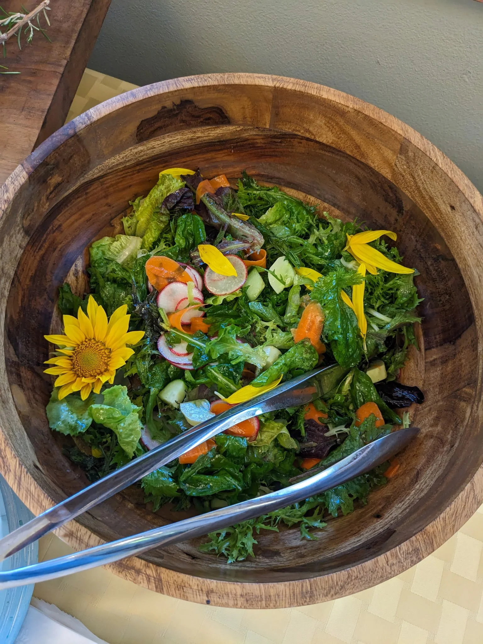 Mixed green salad with radishes, carrots, and edible flowers in a wooden bowl with salad tongs.