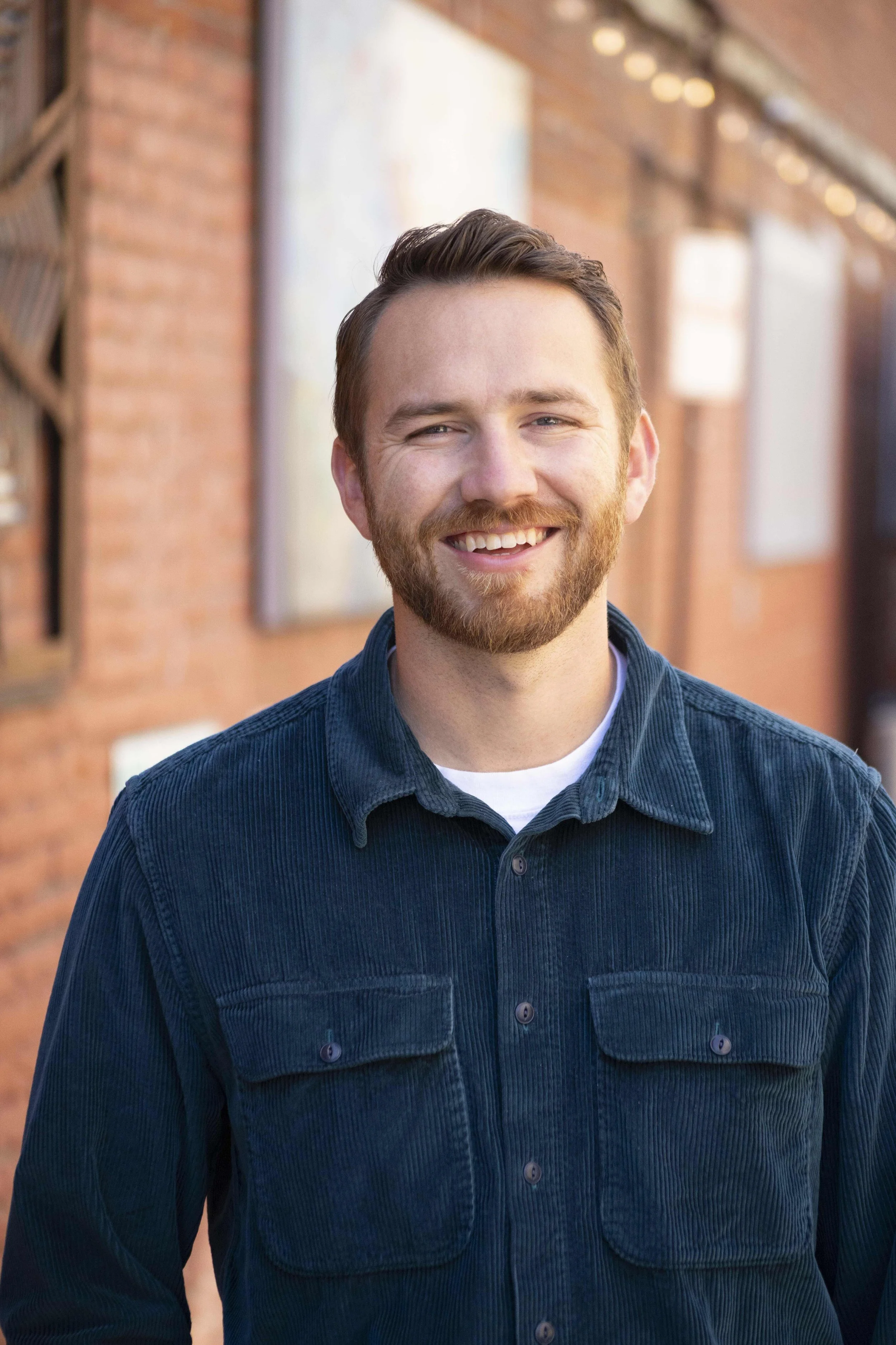 A smiling man wearing a gray Patagonia hat and a bright red Arc'teryx jacket stands outdoors with mountains and a cloudy sky in the background.