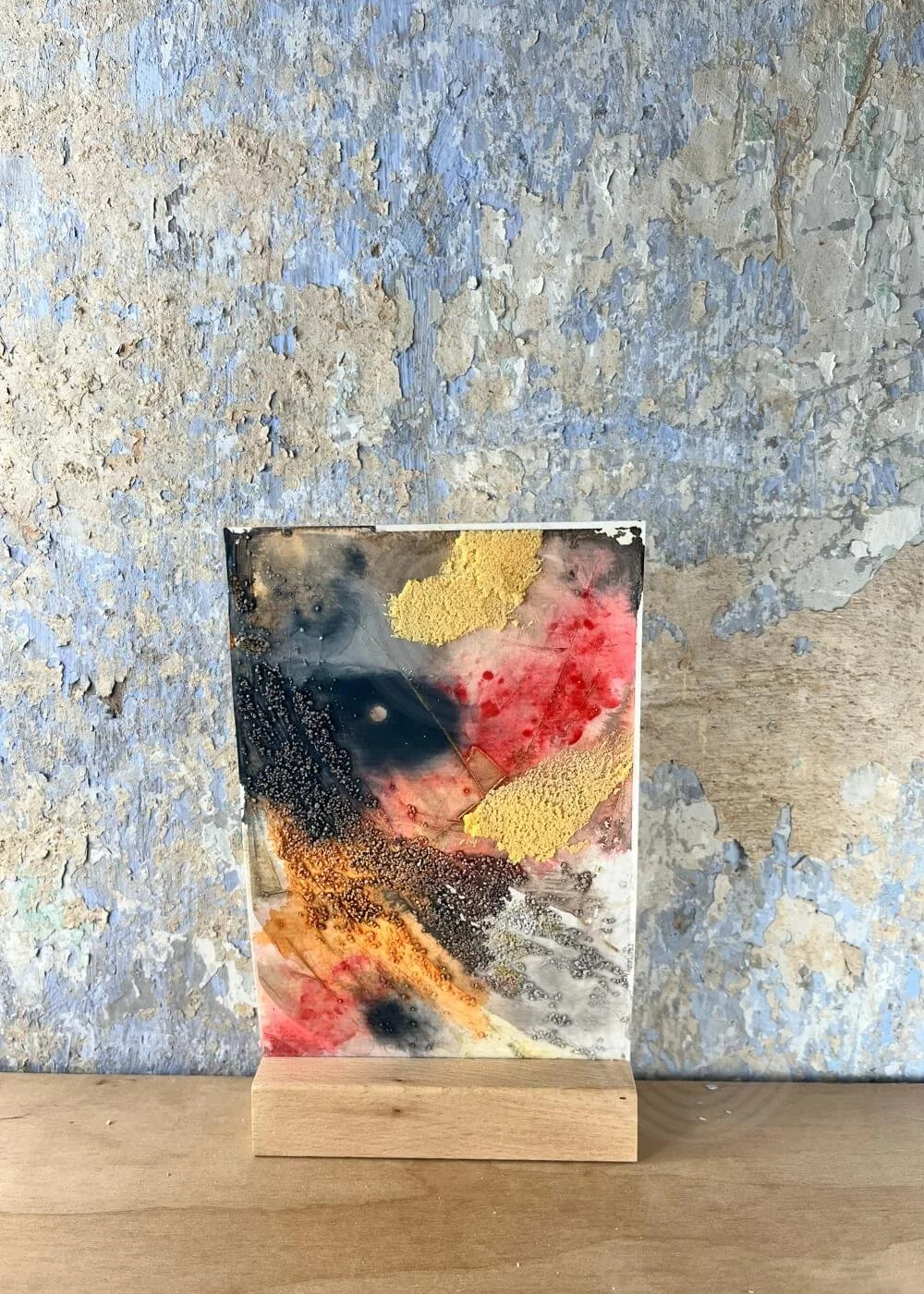 An abstract artwork stands softly against a weathered wall — golden textures and muted reds drift into grey-blue calm, like embers dissolving into dawn.