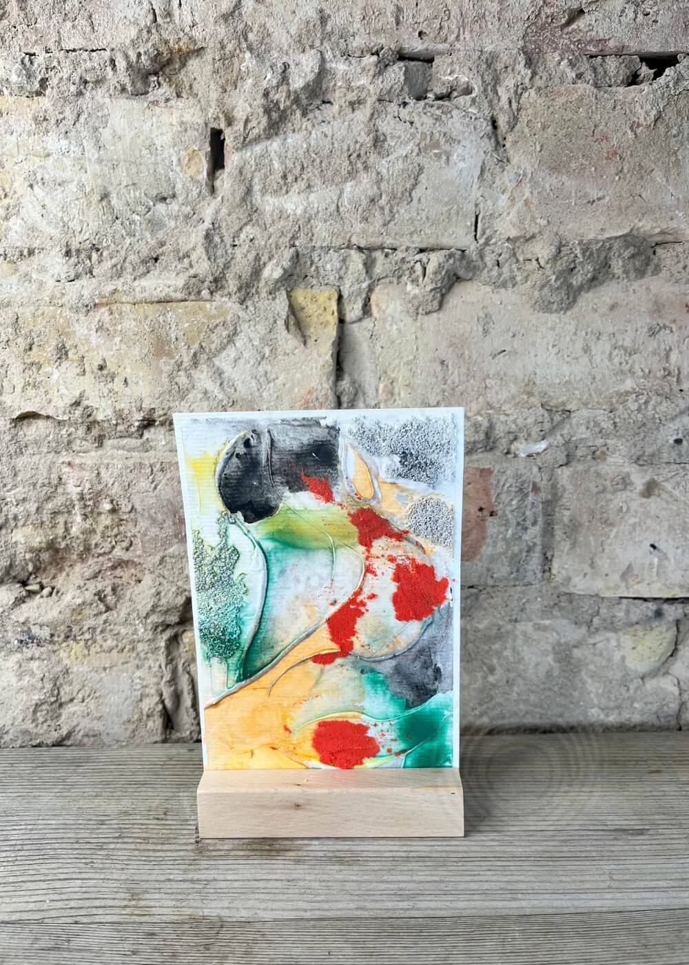 Abstract artwork Risen through fire displayed on a wooden stand against a rustic stone wall, with vivid red, green, and orange hues layered in textured patterns.