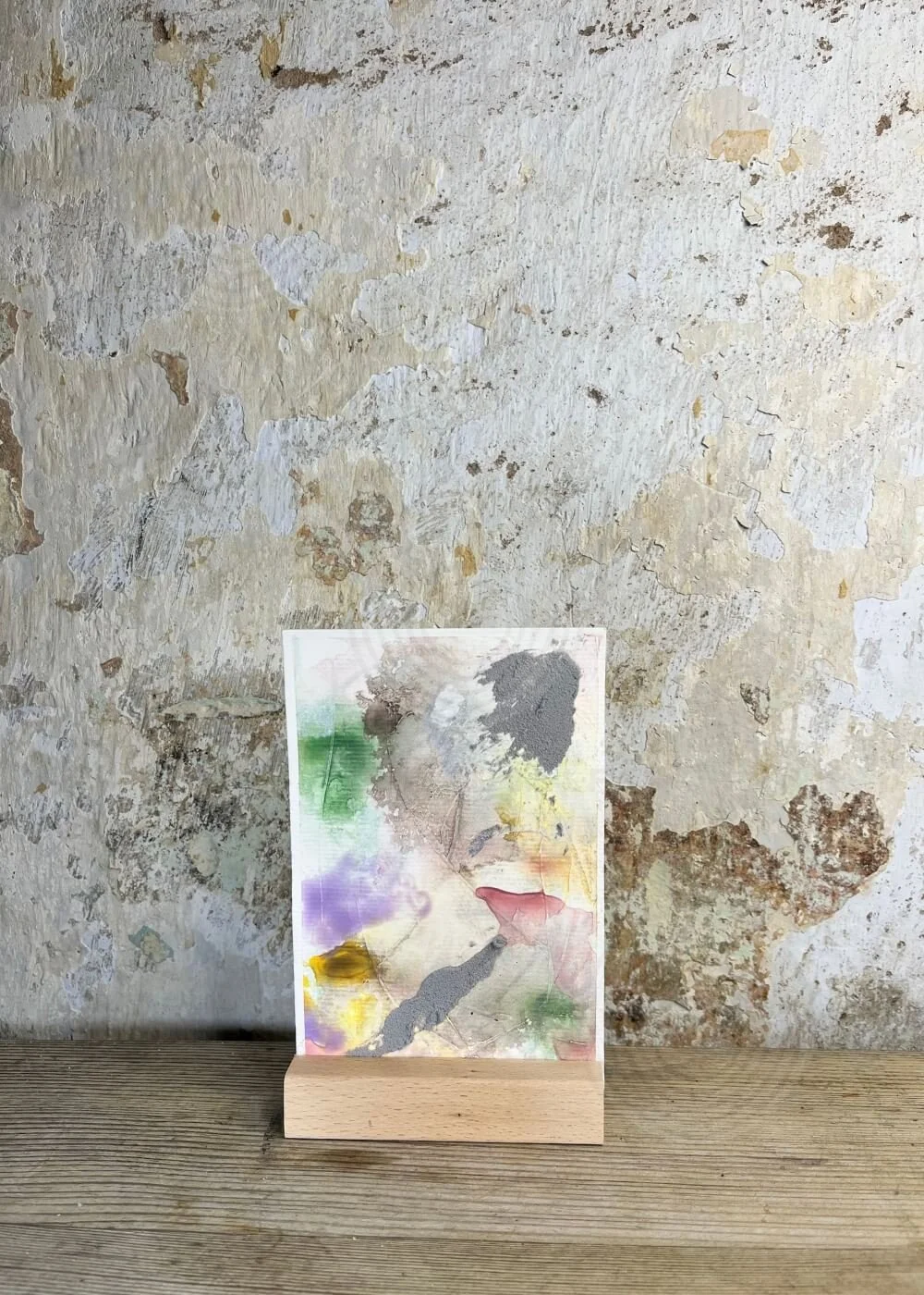 Abstract mixed-media artwork "Between the Seconds No. 9" in soft pastel shades of green, purple, yellow, and grey, displayed upright on a wooden stand against a textured wall.