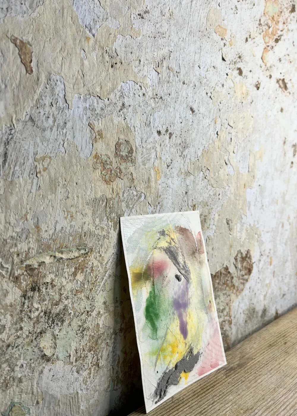 Side view of the small abstract artwork "Between the Seconds No. 1"  with textured layers in green, yellow, purple and grey, leaning against a rustic wall.