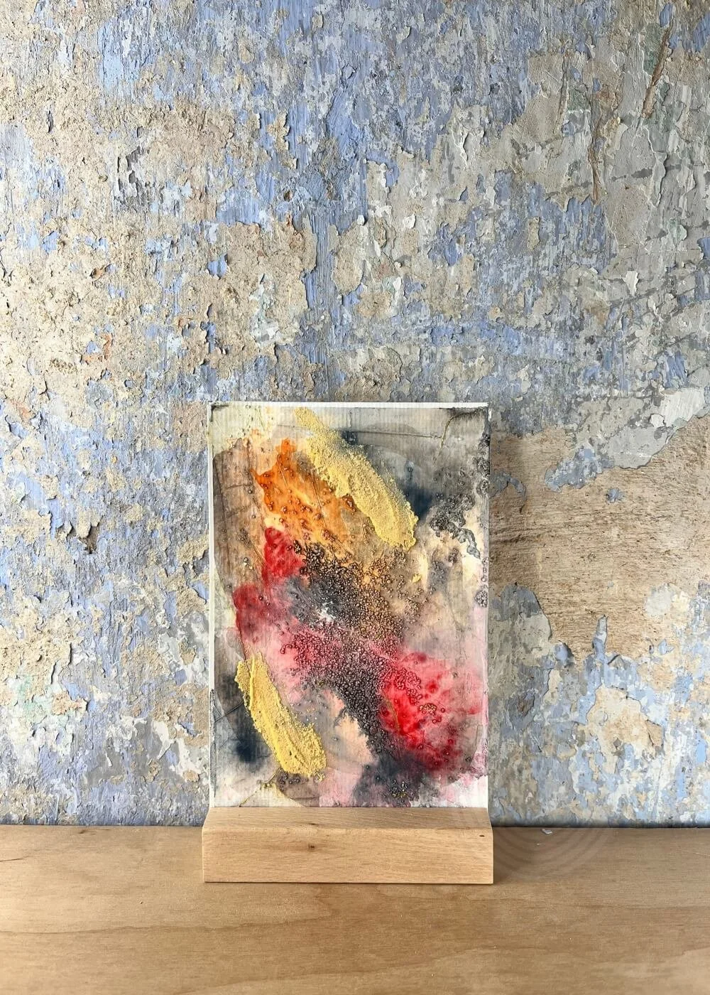 A small abstract artwork resting against a weathered wall — strokes of crimson and ochre meet soft ash-grey, where fire once burned and light quietly remains.