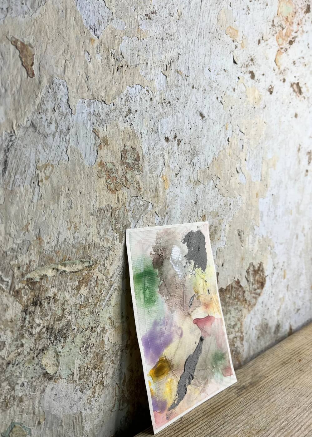 Abstract pastel artwork "Between the Seconds No. 9" with layered textures and earthy tones, leaning against a rustic, weathered wall on a wooden surface.