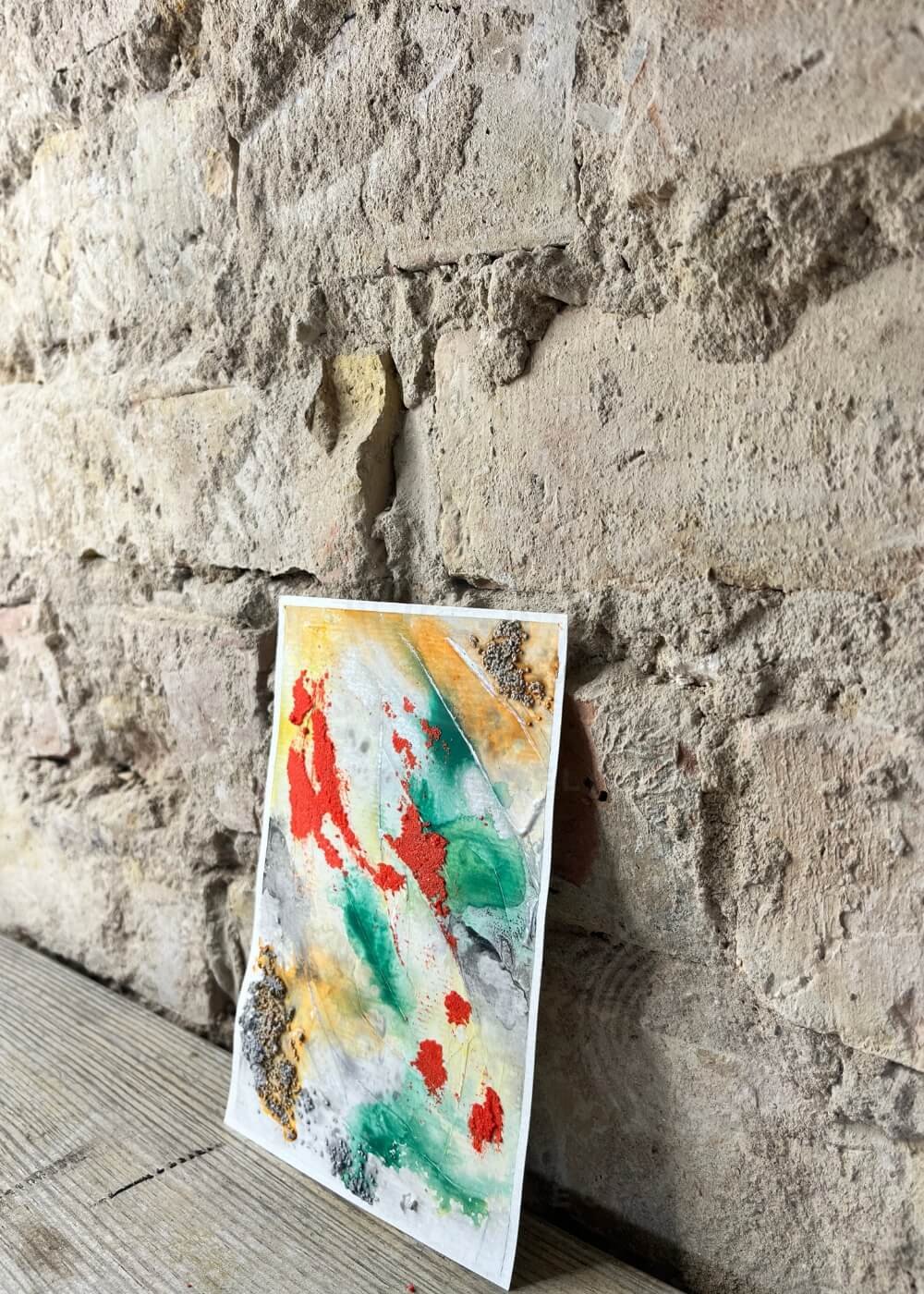 Side angle of abstract healing art piece "Where I Draw the Line" with raised textures in red, green, gold, and gray, leaning against rustic stone wall.