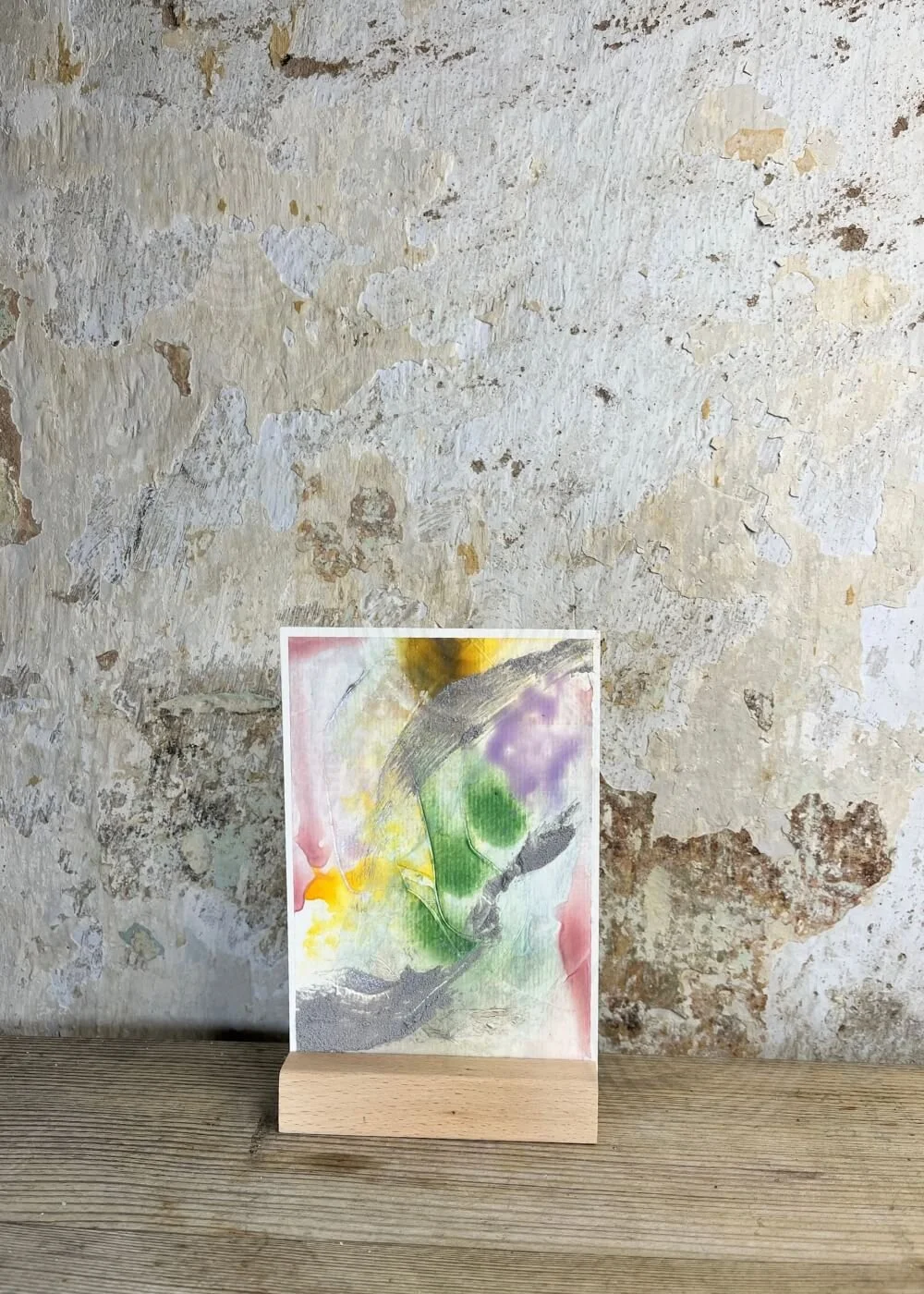 Abstract mixed-media artwork "Between the Seconds No. 8"  with flowing green, yellow, and purple tones, accented by textured grey strokes, displayed on a wooden stand against a rustic wall.