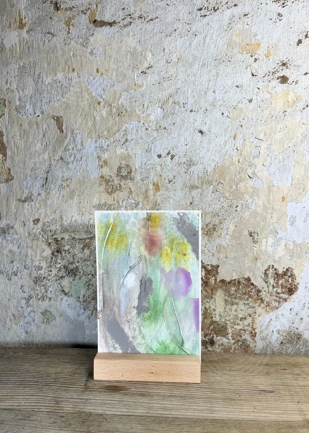 Abstract mixed-media artwork "Between the Seconds No. 2" in soft green, lilac, yellow, and silver tones, displayed upright on a wooden stand against a textured wall.